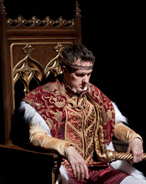King John in ornate royal robes sitting on a throne with his head down pensivelyh