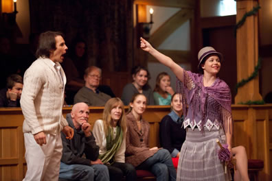 Navarre, in white sweater and pants, reacts to Rosaline, in purple shaw and flapper hat and dress, revealing her love token,  with members of the audience watching behind 