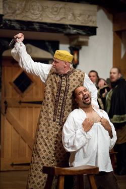 Shylock in long Venitian gown and yellow cap with upraised knife behind Antonio baring his chest