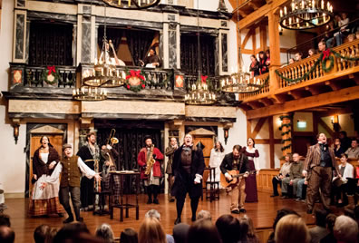 In a theater decked in wreaths and evergreen garland, the cast sings and plays instruments under chandeliers with the audience on three sides. 