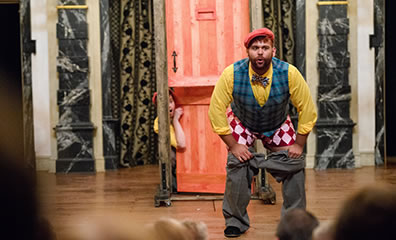 Production photo by Marek K Photography of Dromio of Ephesus with his pants pulled down toward the door that Dromio of Syracuse is peering through.
