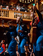 The three wear blue jumpsuits with red trim, and the audience of the wooden Blackfriars is sittein on the stage and in the galleries in the background.