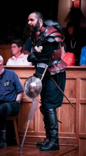 Richard with red armor except a black linkage of plates protruding up the spine, blue pants, a sword at his belt, and a crutch braced to his left hand with a small shield on it.