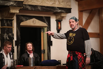 Production photo of Falstaff pointing as he talks, with Gadshill in a sling and Peto with a bloody bandage around her neck sitting at a table behind him. 