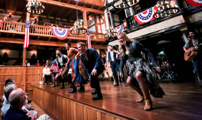 The cast at the front of the stage, legs bent, left leg forward as they sing to the audience, a guitarist, fiddler, standup bass, and drums in the background, bunting hanging from the wood theater walls