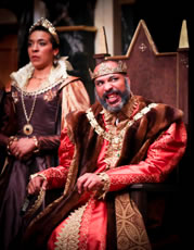 Lear sitting on throne, with gold crown, brown fur robe, gold chain, red shirt and gown, speaking in anger as Goneril in the background, wearing a purple Jacobean dress with gold sleeves, gold medalina, gold tiara, and upturned collar.