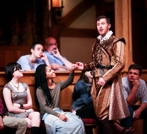 Edmund in brown striped Jacobean knee pants, shirt, and cape, brown vest, and white collar holds the hand of a woman sitting on the gallant stools.