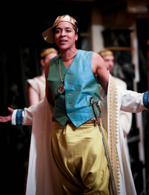Morroco with robe draped over outstretched arms, gold baseball capped turned sidewise, blue vest, gold "S" necklace, gold pants, and silver scimitar through fabric belt.