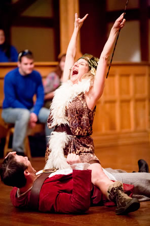 Crocale in a leopard jumper and white boa straddles Tibalt lying on the stage and wearing a red uniform coat and striped sailors pants; Crocale is in triumph, hands upraised, head tilted back, eyes closed, and yelling. Audience members sit in the background.