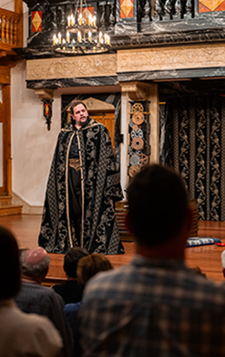 In photo taken from within the audience, Blake Henri stands on stage wearing a silver embroadedred black cloak the exact design of the curtain at the bak of the stage behind him. Also visible are the gear-covered pillars, the bottome of the theater's balcon in marbalized wood, and a chandelier.