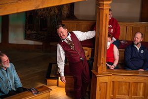 Justin MCombs as Sir Andrew Aguecheek in matching red patterend vest and pants and tie with white shirt hangs out from a wood post among sitting patrons at the back of the playhouse's seating.