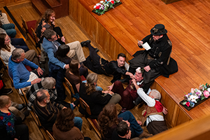 Photo taken from above, black-suited Malvolio sits on the edge of the stage reading a letter in one hand and in the other hand dangles a flower over Aguecheek sitting on the floor in front of him as from eather side, Fabian and Toby Belch are lying on the floor holding extended fingers under Aguecheek's nose in the narrow space between the stage and the feet of  front row patrons. On either side of the stage are trays of flowers.