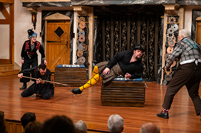 Malvolio wearing black shirt, brown kilt, yellow stockings cross-gartered in black, black shoes, and his black hat with goggles lies on a crate as Christopher Joel Onke's Fabian uses a hook to try to capture one of Malvolio's extended legs. Behind Fabian, Angela Iannone as Maria in black pant suit with red flower collar, blue hair and tiny top hat yells, and to the right Christopher Seiler as Sir Toby Belch in short gray plaid jacket and brown pants accosts Malvolio. In the background is the door, gear-covered columns, and silver-embroidered black curtain of the wood stage, and in front the tops of watching patrons heads.