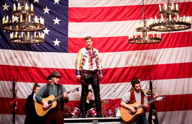 Jackson in yellow Victorian-era uniform jacket, bloody white t-shirt, black leatherpants and boots stands on the desk in front of a drum set with two guitarists on the side (both wearing country vests, one in hat), U.S. flag in the background and old chandeliers overhead