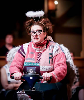 The Storyteller, with her hair tied up in two bunches like mouse ears, wears a pink-dominant boldly patterned sweater, black horned-rim glasses, and furry white hallow and wings as she sits on her scooter.