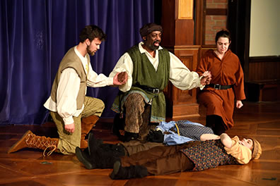 Production photo Will Kirk Photography of Belarius between Arviragus and Guiderius, kneeling and holding hands behind the two bodies on the stage
