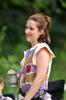 Hal in a vest with purple, gold, and red patches intended to look like chain mail with a black sword belt around her waist holds a pewter mug in her hand. Trees are in the background.