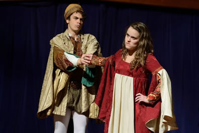 Petruchio in renaissance pants (green) and white hose with brown brocaded jacket and gold brocaded cloak and aht holds on to Kate's right arm while she frowns with tongue in cheek and left hand on hip. She's wearing a red dress with a white panel on front and long draping white sleeves. 