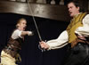 Mercutio in leather jerkin thrusts his sword with ayell as Tybalt in yellow fest parries Mercutio's thrust with his sword while his left hand sits bent at the hip.