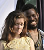 Phoebe with windswept brunette hair and wearing a yellow dress has a frown in her expression while behind her, Sylvius with Bohemian shirt and serape vest smiles broadly at her.