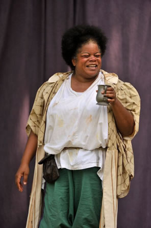 Falstaff, with white puffy shirt, green pants, black coin purse hanging on her string belt, and a tapestry like cloak holds up a pewter mug