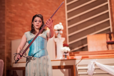 Aspatia in a filly blue patterned cream dress and pastel blue corset strokes a long sword she is holding in her hands. In the background is the sancuary of the church