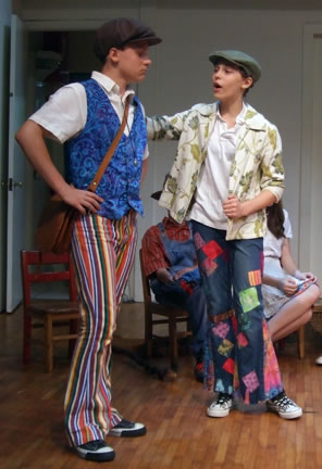 Orlando in dog cap, white shirt, blue paisely vest, multicolor striped pants, and brown leather manbag across his shoulder, hands on hips, Rosalind also in dog cap, white shirt, leavy tan leisure jacket, blue jeans with colorful patches, other actors sitting on chairs in the background.