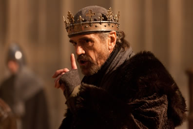 Henry IV, with fur cap and fingerless gloves, simple gold crown over a skull cap, points a finger up as he speaks with a threatening look