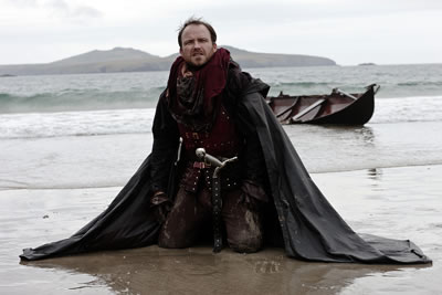 Bolingbroke in waistcost armor, sword in hilt, cloak about him, neals on a muddy beach with a boat in the surf behind him