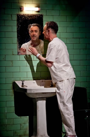 Alan Cumming at a sink and wearing hospital patient shirt and pants looks into the mirror