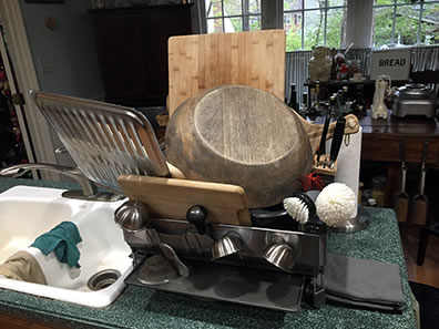 Photo of dish drainer filled with wood cutting boards and bowl, little silver bowls hanging off the side, and a broiler pan dangling off the edge over the sink.