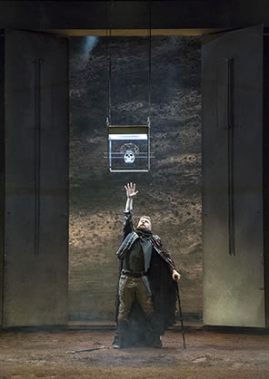 Production photo of Richard standing under the crowned skull in the glass box suspended from the ceiling. Richard is reaching up as high s he can with his right arm while his left arm leans on a crutch (the other crutch is at his feet on the dirt floor.