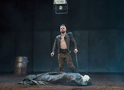 Production photo of Richard, in black unbuttoned Renaissance jacket and bround pants, leaning on his two crutches, standing by the shrouded body of Henry VI. The floor is dirt, a barrel is in the background, and a skull is suspended in a glass box hanging from the ceiling.