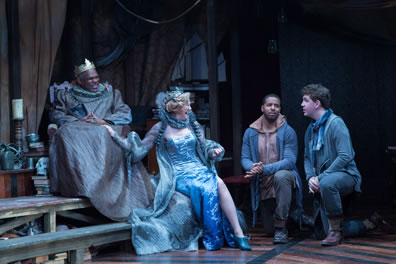 In a blue tinted light, Claudius in a blue satin robe and large gold crown sits in a chair with sitting next to him, her hand on his knee, is Gertrude in a shimmering blue split-sided evening own and cape, both wearing blue ruff collars. Rosencrantz and Guildenstern are on one knee across from them. 