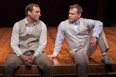 Valentine and Proteus dressed similarly in tan shirts and pants with matching vests sit on the edge of the stage talking, Proteus with his left foot upon on the stage and arm resting on his knee.