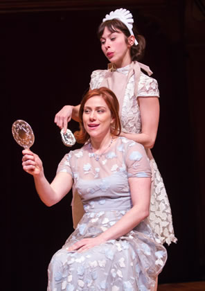 Julia sitting in a long mauve gown with white patterns holds a mirror up while behind her Lucetta in white lace dress, apron, and maid's crown brushes Julia's hair
