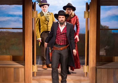 Gunslinging Petruchio in red vest and black overcoat and cowboy hat leas Kate in red riding coat and cowboy hat and Grumio in yellow buttler's tailes and feathered bowler through the saloon doors