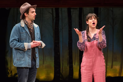 Silvius in fodora hat, jeans jacket over dark olive vest, patterned scarf, and jeans with watches Phoebe talking with her hands up and wearing pink overalls and a pink and purple pattereed shirt. In the background is a backdrop of trees.