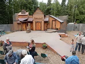 Photograph of the outdoor Fairbanks Shakespeare Theatre stage, with the wood facade Merry Wives set, ramps into the audience in chairs on the ground and platforms. Trees in the background
