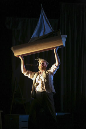 Prospero, in white shirt and rustic brown vest, bearded and wearing his thorn of twigs and a large necklace holds over his head a small wooden sailboat.