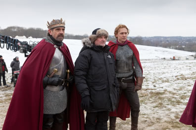 Irons and Hiddleston, dressed in medieval armor with red cloaks, stand on either side of Eyre in a parka and winter hat with snow on the ground and a camera boom in the background