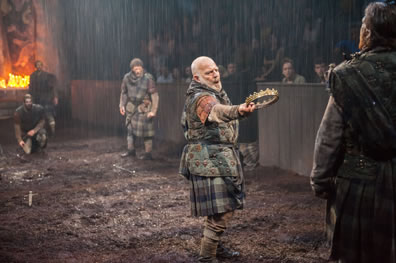 Duncan, in muted tartan kilt, holds out his crown to an opposing warrior with his back to us as other warriors watch; you can see the rain following, candles from one end of the stage in the background, and the audience sitting behind walls.