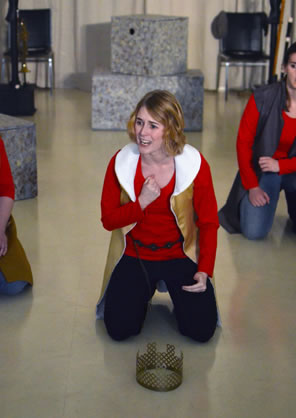 Richard II kneels on the ground with the crown on the floor before her as she pounds her chest, other members of the cast kneeling behind her
