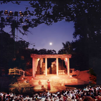 An audience watches a play on an outdoor stage under a moonlit sky
