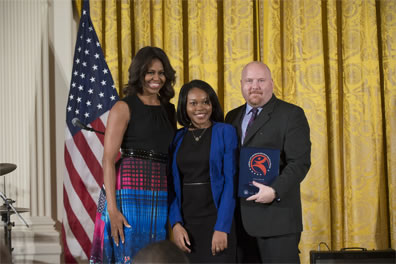 Michelle Obama in black tank top and red, blue dress with checked pattern and flowers, Sendie Brunard in black dress and blue sweater,, Pound in dark gray suit holding award. American flag behind first lady, gold curtain across the back to the right, drum set to the left.