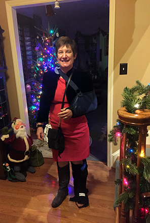 Photo of Sarah in nice red dress, braced left arm in sling, left foot in orthopedic boot with Christmas tree and Santa in background, lit-garland stairway railing at forefront.