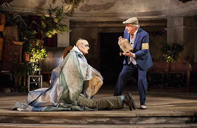 Production photo of Caliban wearin gaberdine sitting on top of Trinculo as Stephano holds before him a bottle.