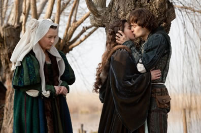 Nuse in green renaissance gown and white headdress with her head down and hands in worry position stands beside a kissing Juliet in black cloak and Romeo in blue