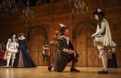 On a stage with an oak wood screen, two doors with greenery over their arches, candelabra and candle chandeliers, in the close right is Viola in white courtier outfit with cap and long red hair, Orsino in olive green elizabethen wear kneeling, in the distance is Sebastian looking just like Viola and Olivia in black dress with puple seem, and Feste leaning against the back wall in red, green, and white smock and green tights and bootsOn a stage with an oak wood screen, two doors with greenery over their arches, candelabra and candle chandeliers, in the close right is Viola in white courtier outfit with cap and long red hair, Orsino in olive green elizabethen wear kneeling, in the distance is Sebastian looking just like Viola and Olivia in black dress with puple seem, and Feste leaning against the back wall in red, green, and white smock and green tights and boots