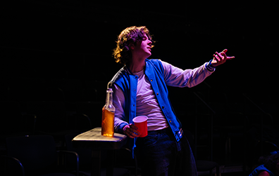 SIG-This_Ends_In_Blood-Duncan-5 Duncan, wearing a blue and white letter jacket over a whit sweat shirt, gestureses with his left hand while holding a red cup in his right hand, his right elbow leaning on a counter on which is placed a long-neck bear.